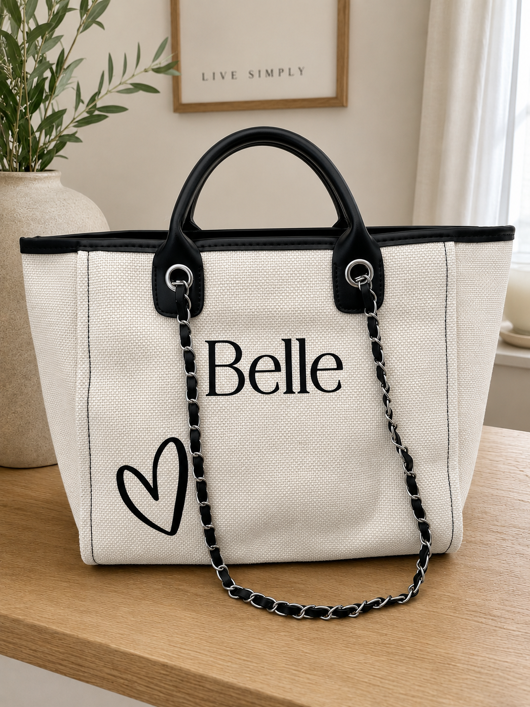 A cream canvas tote bag with black trim, black top handles, a black chain strap, and the word 'Belle' printed in black alongside a heart outline, displayed on a wooden table in a home setting.